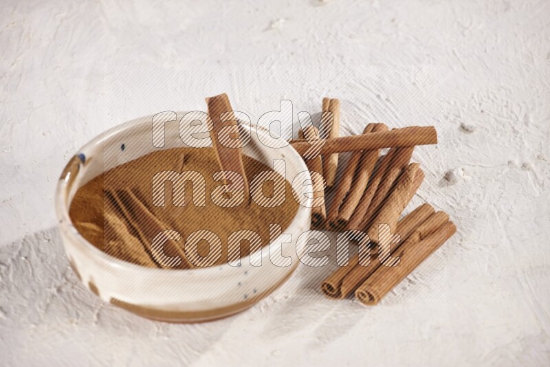Ceramic bowl full of cinnamon powder with cinnamon sticks on the side on white background