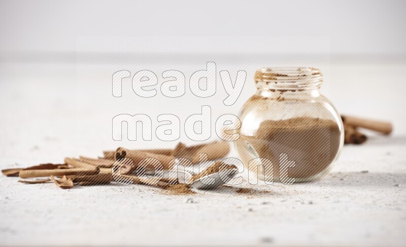 Herbal glass jar full cinnamon powder and a metal spoon surrounded by cinnamon sticks on a white background