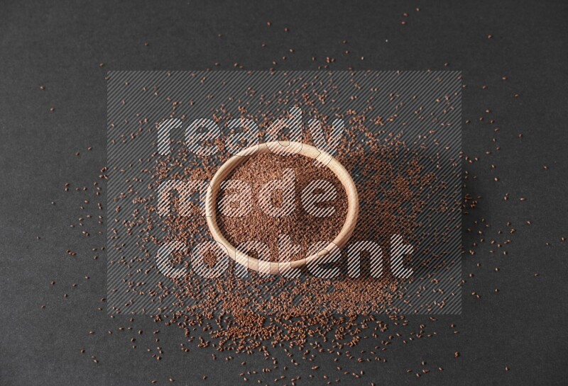 A wooden bowl full of garden cress seeds surrounded by seeds on a black flooring