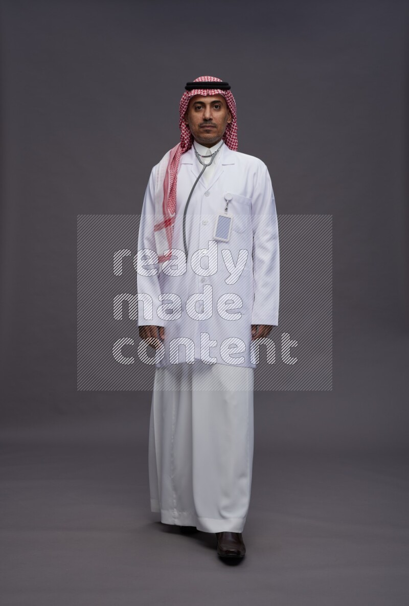 Saudi man wearing thob with lab coat and shomag with pocket employee badge with stethoscope standing interacting with the camera on gray background