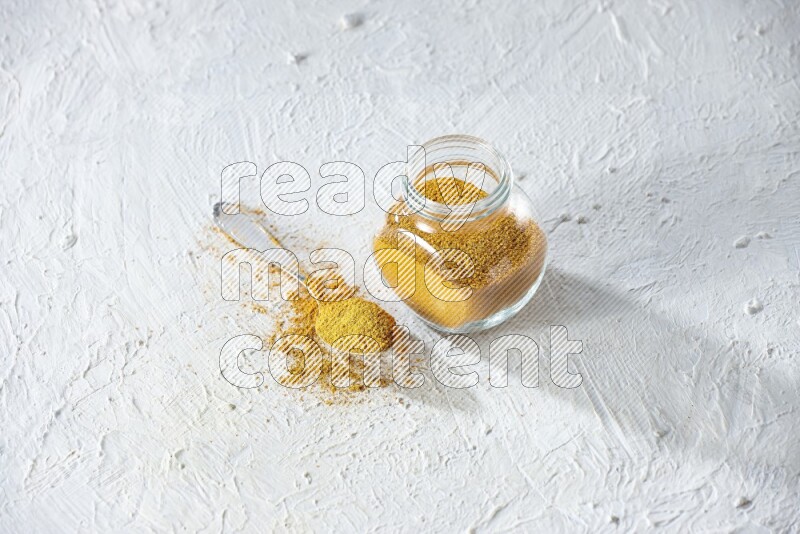 A glass spice jar and metal spoon full of turmeric powder on textured white flooring