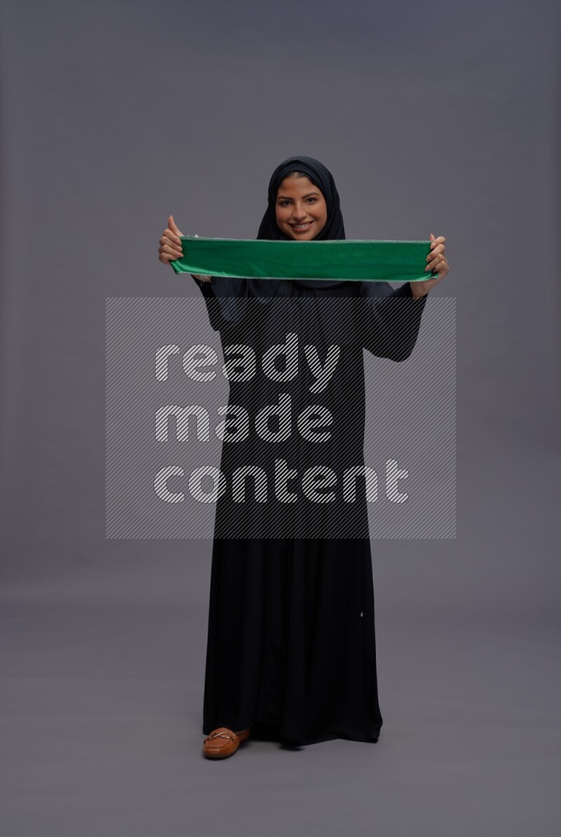 Saudi woman wearing Abaya standing holding Saudi flag on gray background