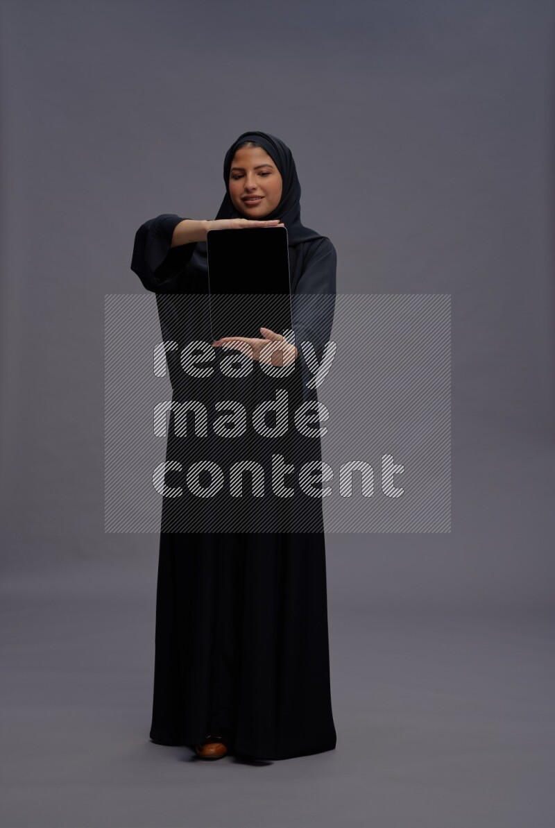 Saudi woman wearing Abaya standing showing tablet to camera on gray background