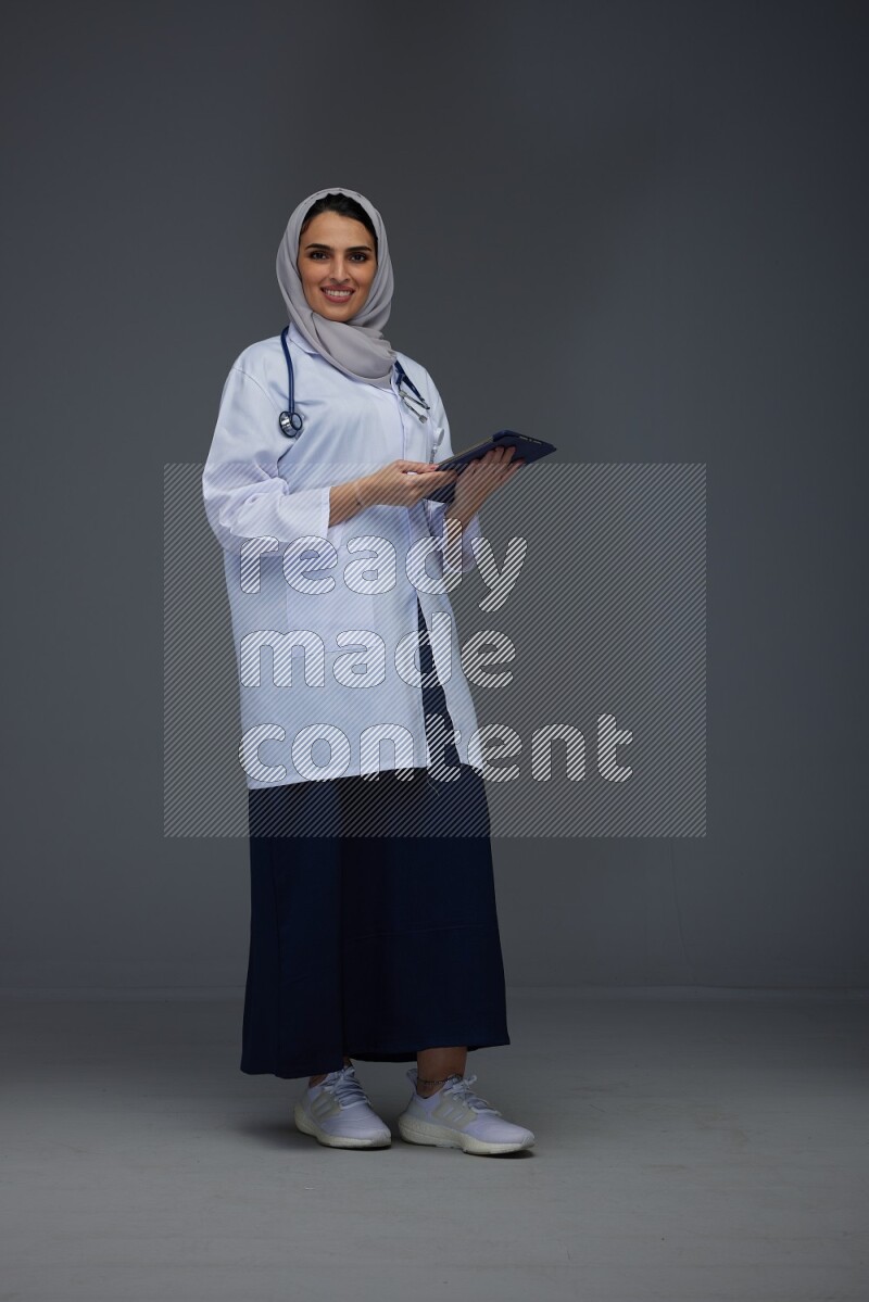 A Saudi doctor wearing a light gray head scarf standing and crossing her hands eye level on a grey background