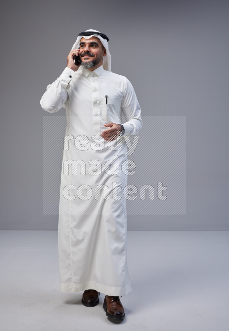 Saudi man Wearing Thob and white Shomag standing talking on phone on Gray background