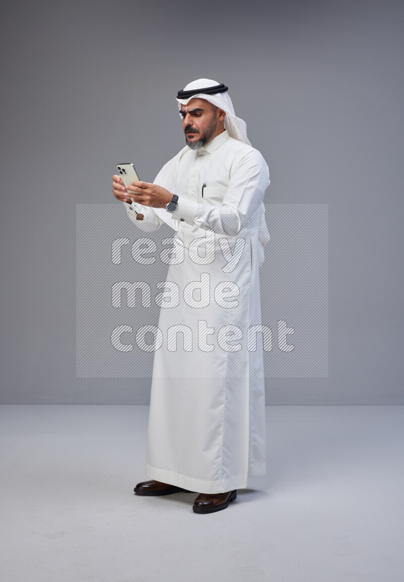 Saudi man Wearing Thob and white Shomag standing texting on phone on Gray background