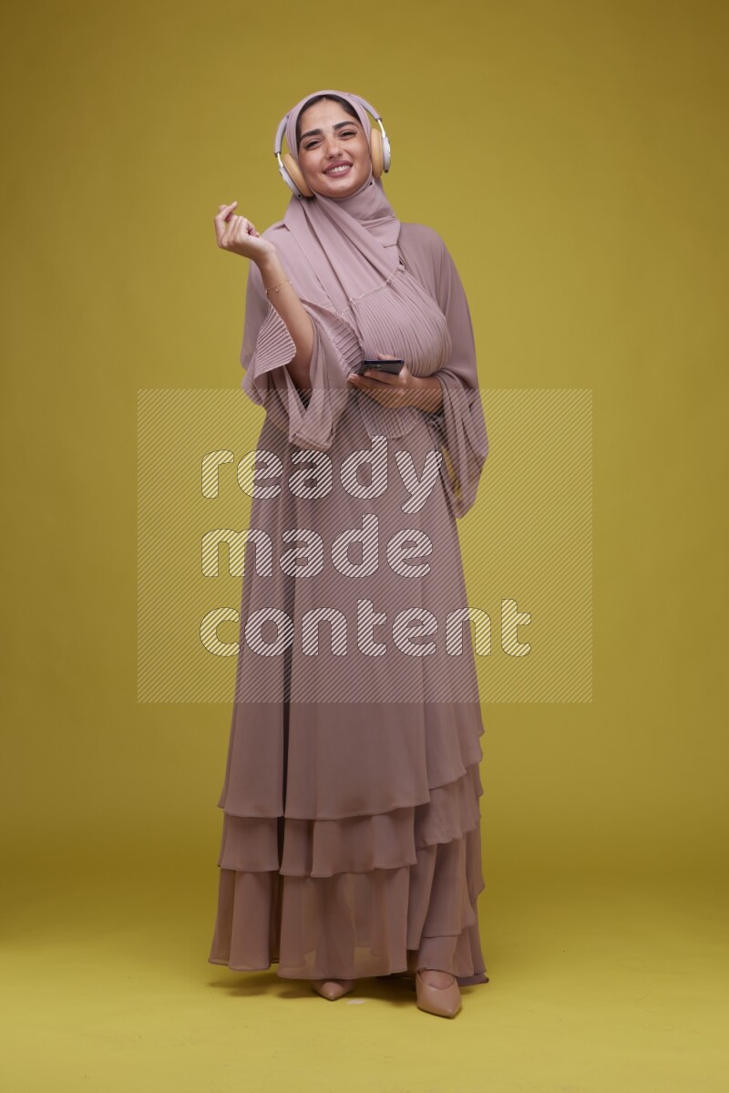 A woman Listening to Music on a Yellow Background wearing Brown Abaya with Hijab