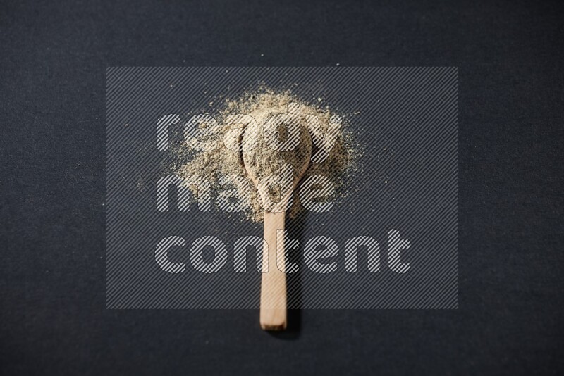 A wooden spoon full of cardamom powder and powder spreaded on black flooring
