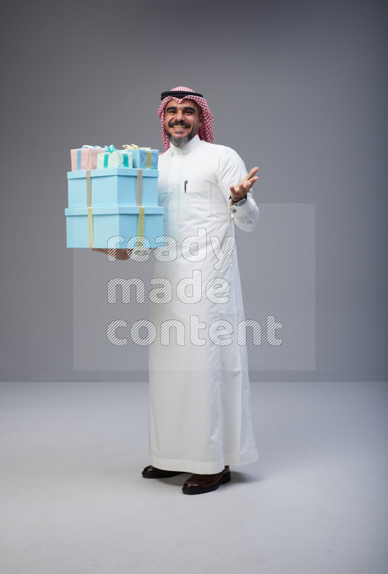 Saudi man Wearing Thob and red Shomag standing holding gift box on Gray background