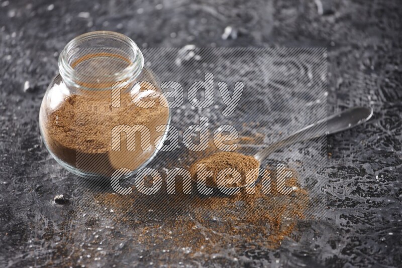Herbal glass jar full of cinnamon powder and a metal spoon full of powder on textured black background