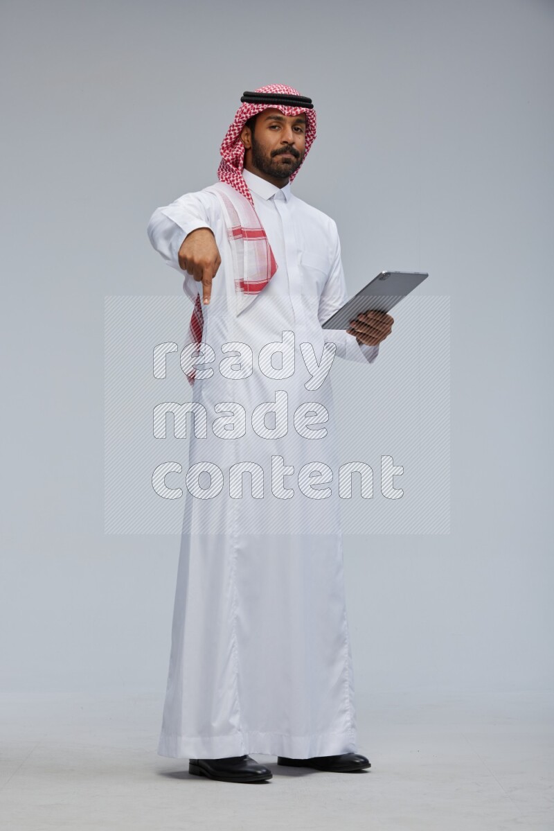 Saudi man Wearing Thob and shomag standing working on tablet on Gray background