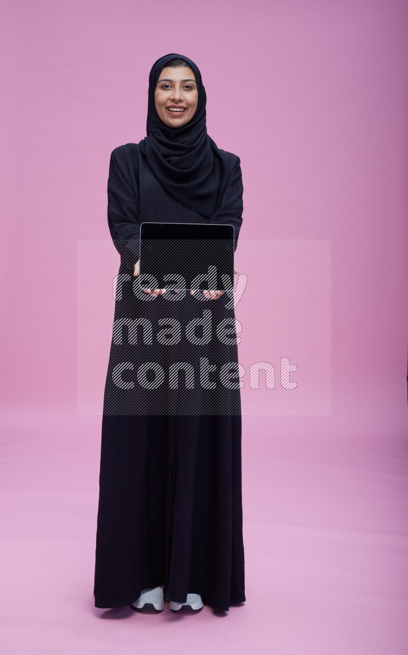 Saudi woman wearing Abaya standing showing tablet to camera on pink background