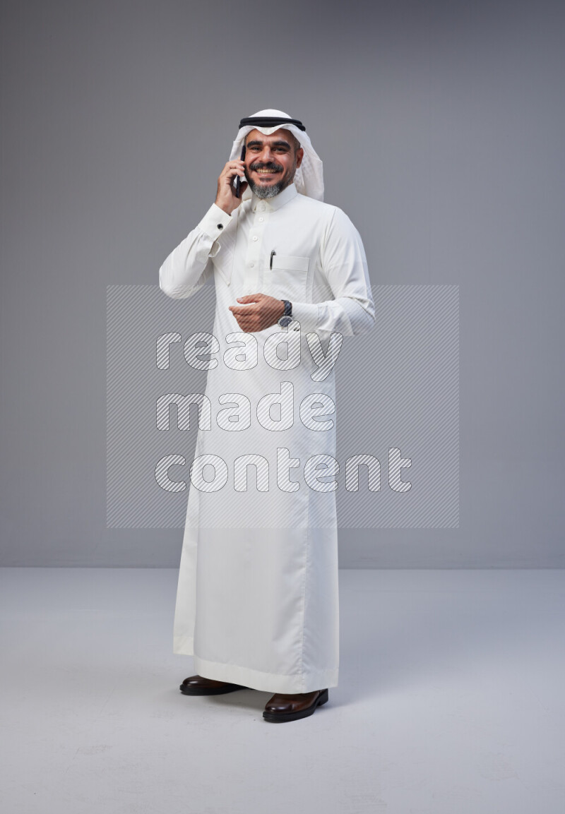 Saudi man Wearing Thob and white Shomag standing talking on phone on Gray background