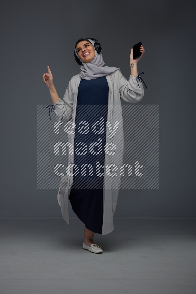 A Saudi woman wearing a light gray Abaya and head scarf standing and listening to music on a grey background