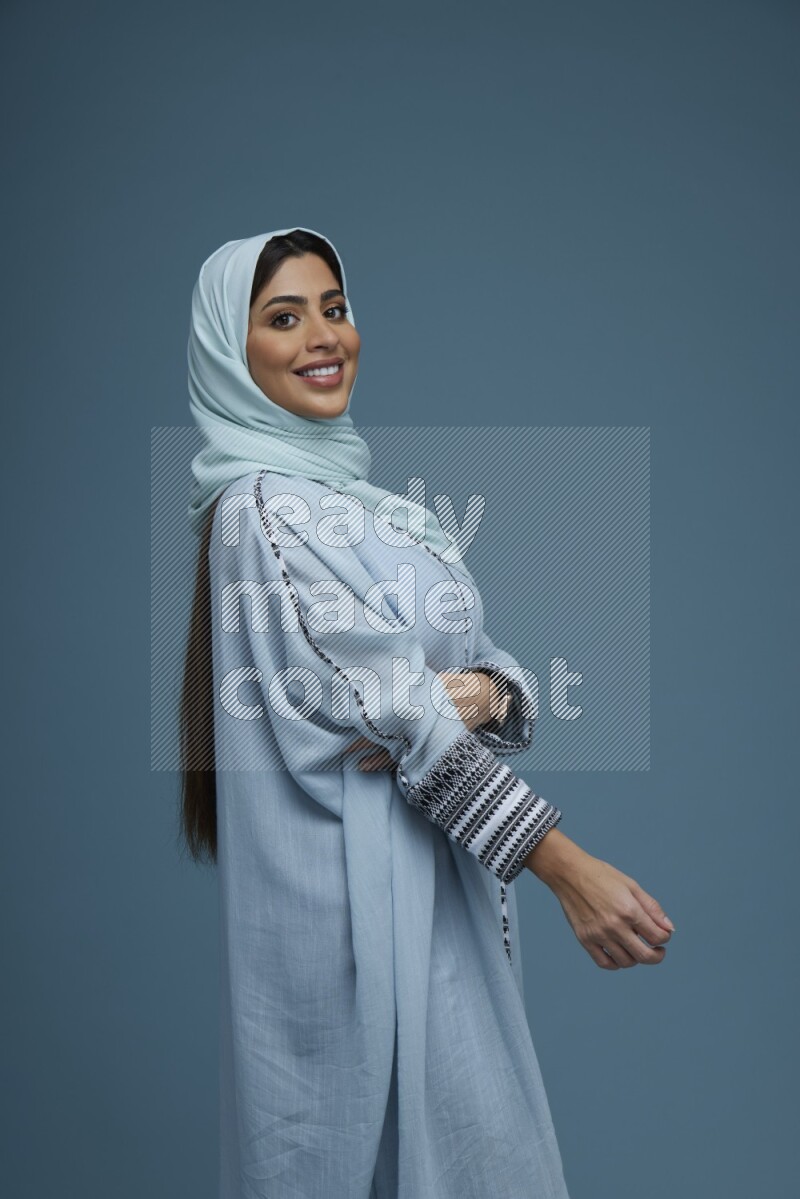 A woman posing in a blue background wearing a blue Abaya with hijab