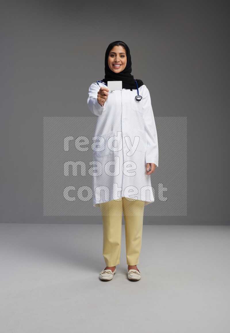 Saudi woman wearing lab coat with stethoscope standing holding ATM card on Gray background