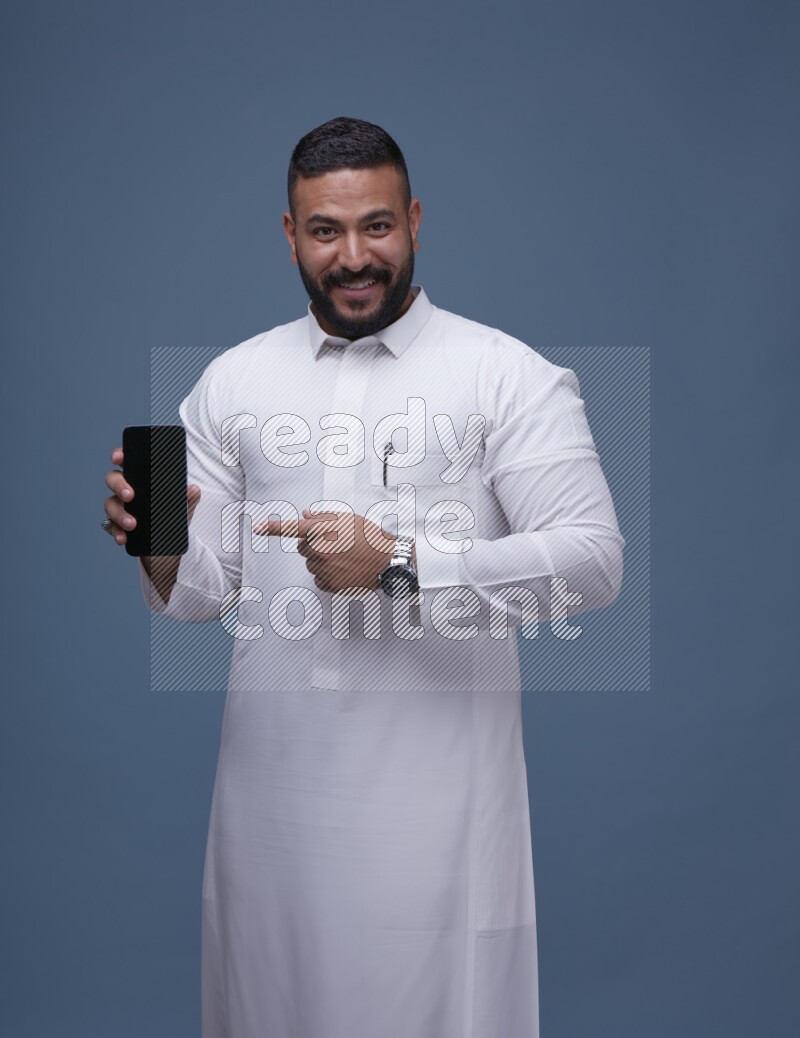 A man Showing his phone in Blue Background wearing Saudi Thob