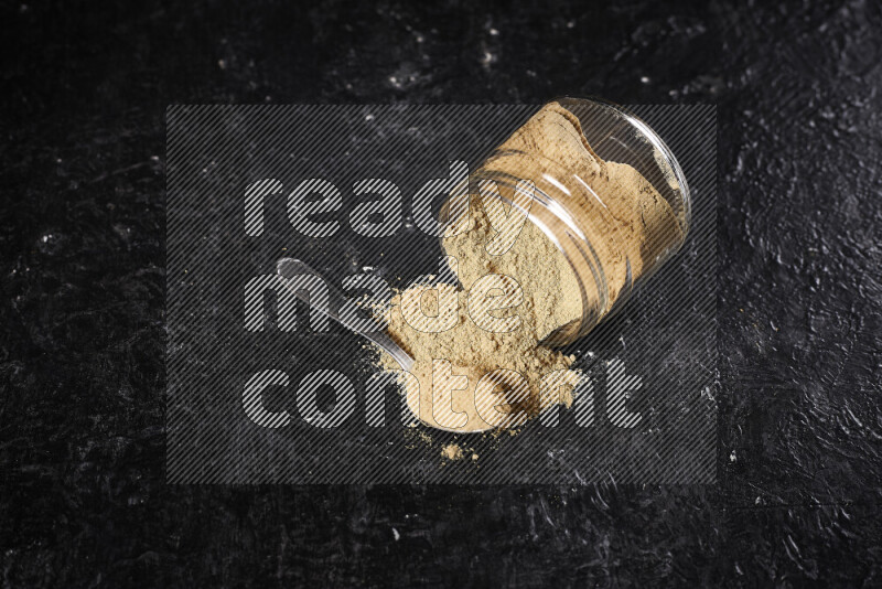 A glass jar full of ground ginger powder flipped with some spilling powder on black background