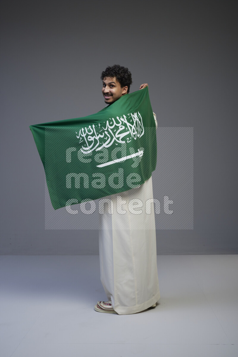 A saudi man standing wearing thob holding big saudi flag on gray background