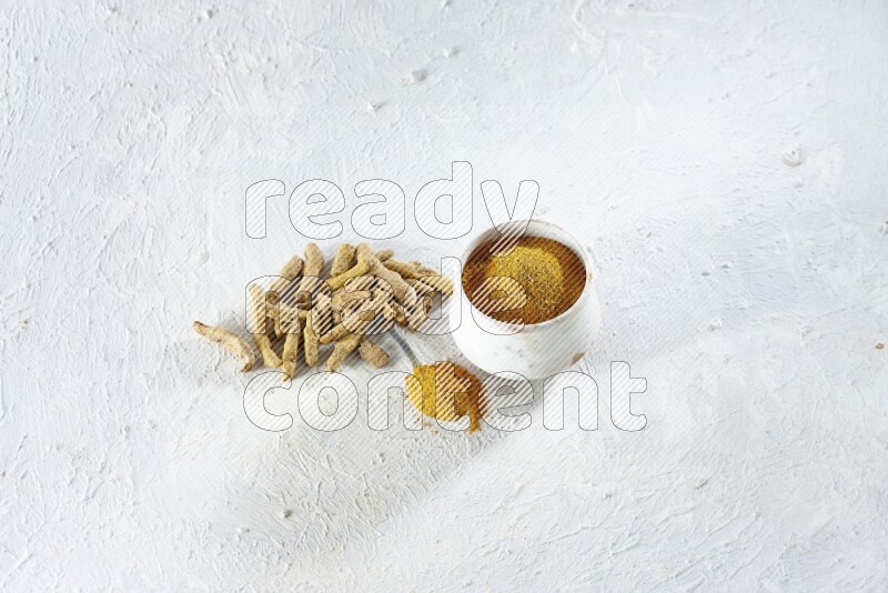 A beige pottery bowl and metal spoon full of turmeric powder and dried turmeric fingers next of them on textured white flooring