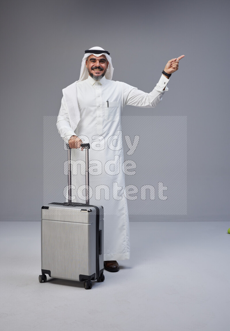 Saudi man wearing Thob and white Shomag standing holding Travel bag on Gray background