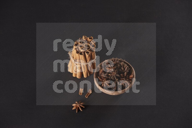 A stacked and bounded cinnamon sticks and a wooden bowl full of star anise on a black background