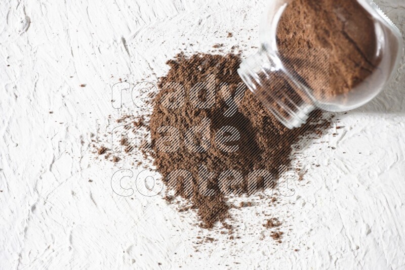 A flipped glass spice jar full of cloves powder and powder came out of it on textured white flooring