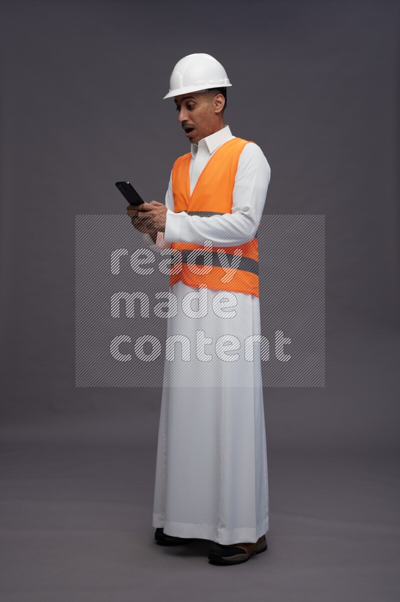 Saudi man wearing thob with engineer vest standing texting on phone on gray background