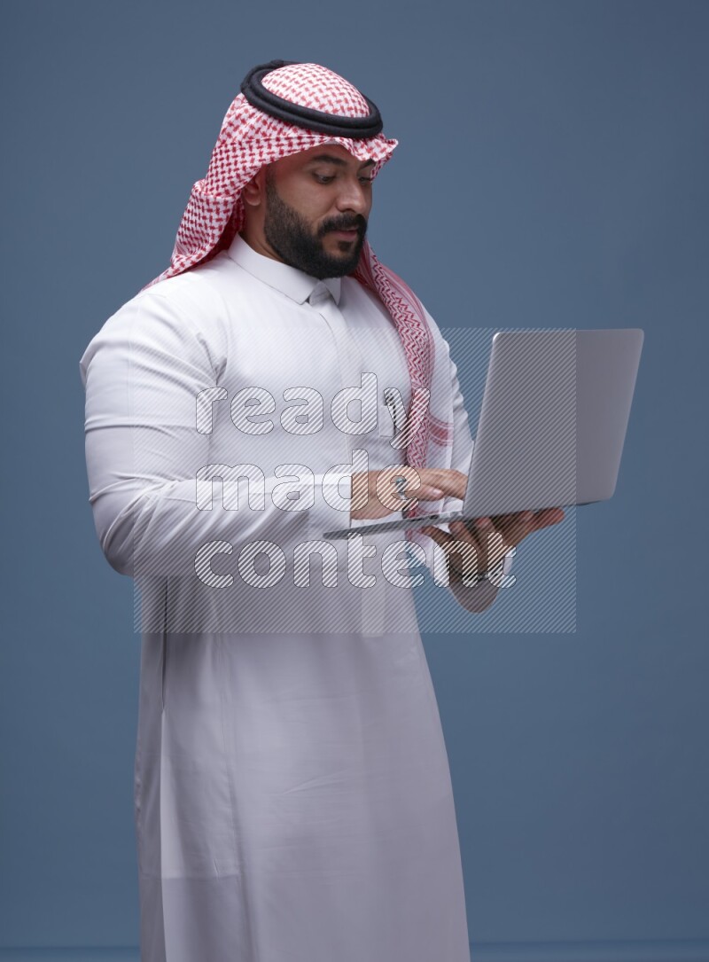 A man standing  with a laptop on Blue Background wearing Saudi Thob and Shomag