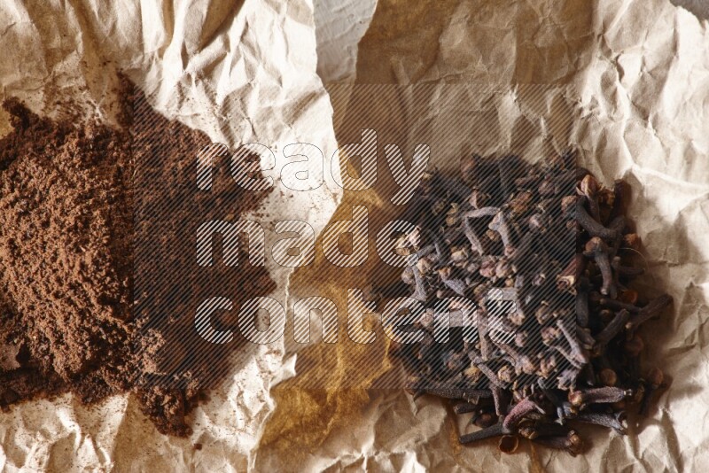 Cloves powder and cloves on 2 crumpled pieces of paper on a textured white flooring