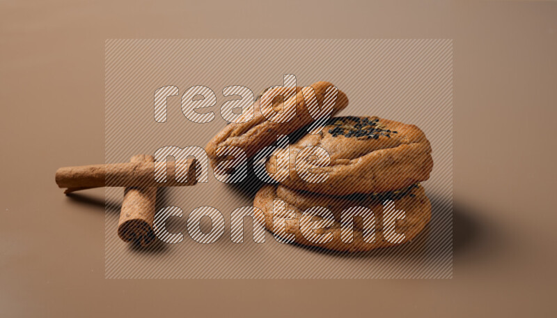 Three chocolate chip cookies beside cinimon sticks on a brown background