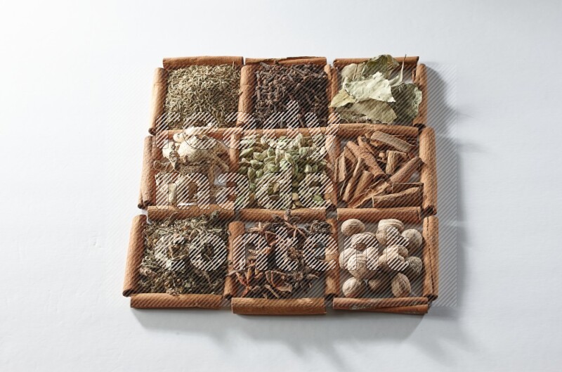 9 squares of cinnamon sticks full of cardamom in the middle surrounded by nutmeg, cinnamon, bay laurel leaves, cloves, cumin, dried ginger, dried basil and star anise on white flooring