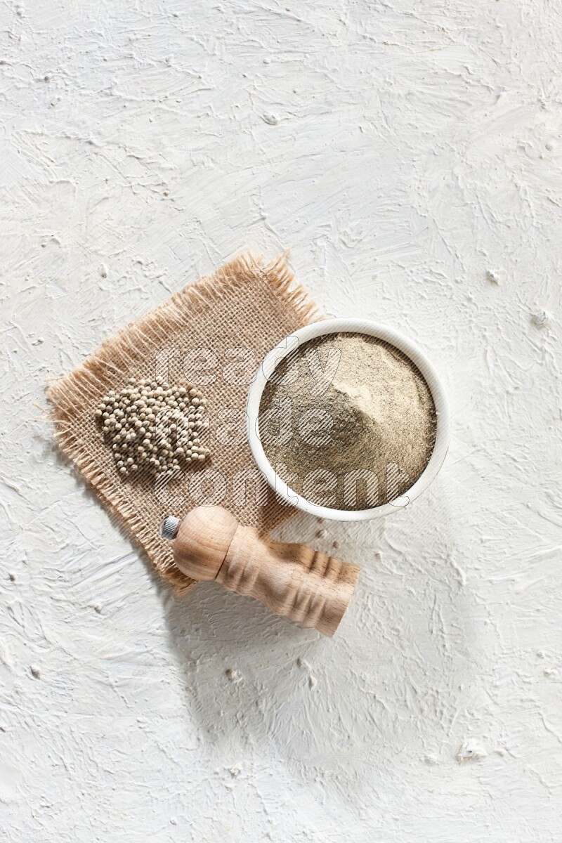 White pottery bowl full of white pepper powder set on a burlap piece of fabric with pepper beads and wooden pepper grinder on textured white flooring