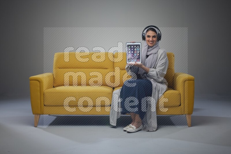 A Saudi woman wearing a light gray Abaya and head scarf sitting on a yellow sofa and using her phone while wearing headphones eye level on a grey background