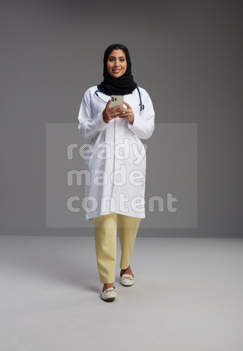 Saudi woman wearing lab coat with stethoscope standing texting on phone on Gray background