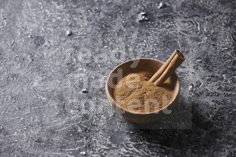 Wooden bowl full of cinnamon powder and a cinnamon stick on a textured black background