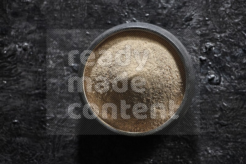 Black pottery bowl full of white pepper powder on textured black flooring