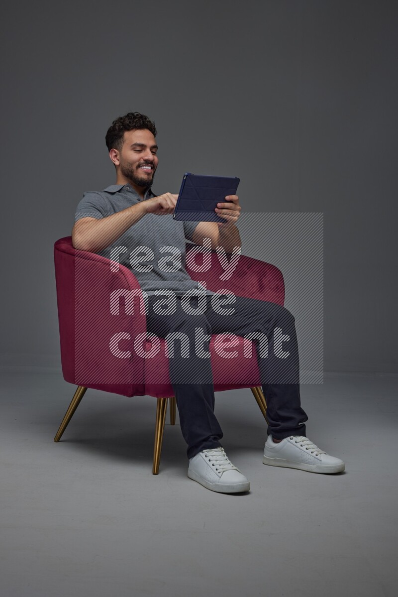 A man wearing casual and using his tablet while sitting on a burgundy chair eye level on a gray background