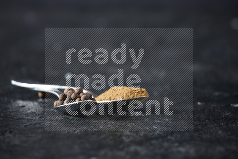2 metal spoons full of allspice powder and whole balls on a textured black flooring