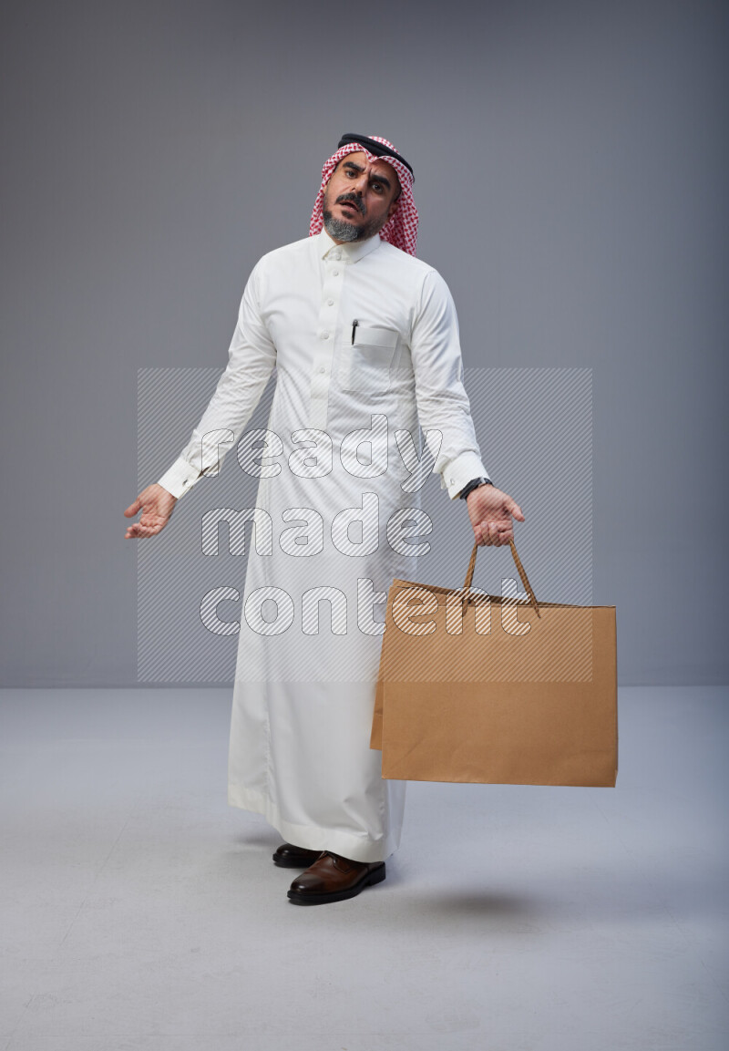 Saudi man Wearing Thob and red Shomag standing holding shopping bag on Gray background
