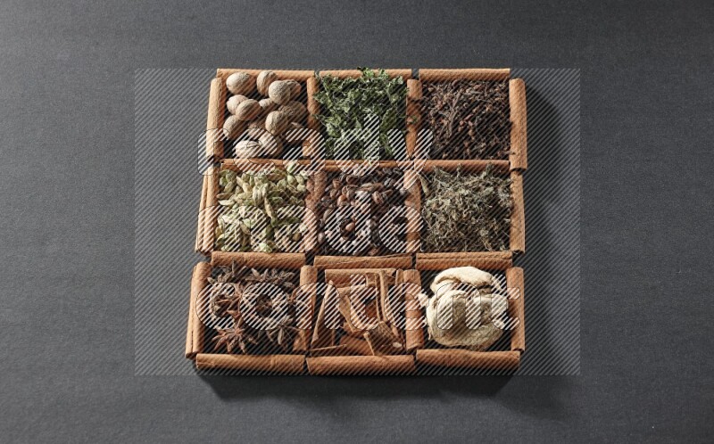9 squares of cinnamon sticks full of coffee beans in the middle surrounded by nutmeg, dried mint, cloves, dried basil, dried ginger, cinnamon, star anise and cardamom on black flooring