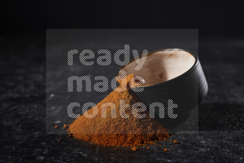 A black pottery bowl full of ground paprika powder with fallen powder from it on black background