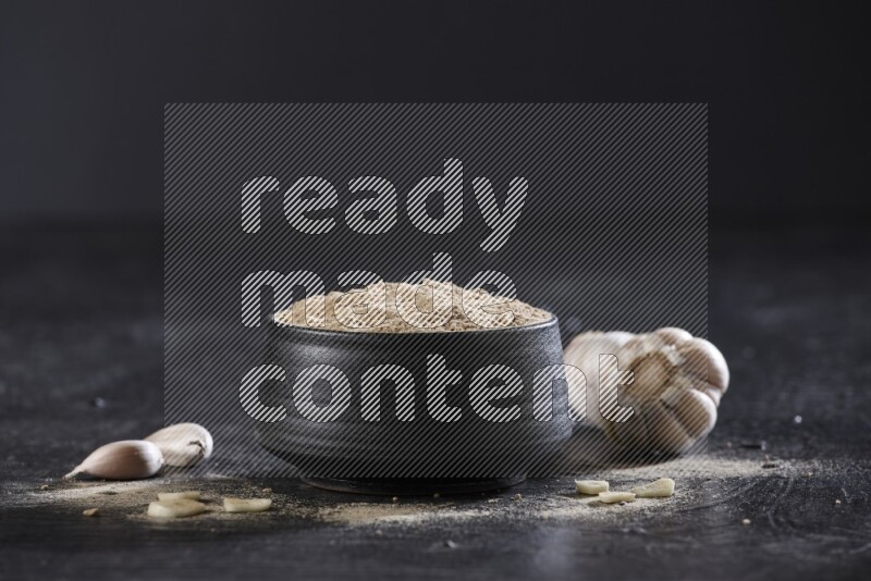A black pottery bowl full of garlic powder with garlic cloves and a whole bulb beside it on a textured black flooring