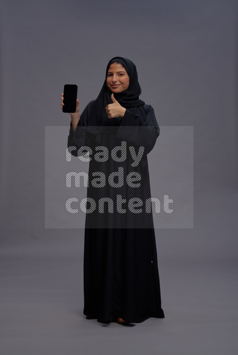 Saudi woman wearing Abaya standing showing phone to camera on gray background