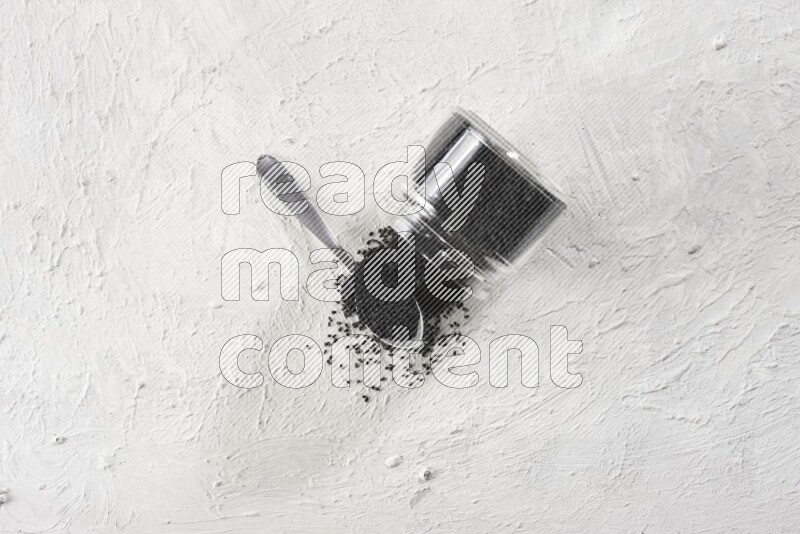 A glass jar and a metal spoon full of black seeds and the jar flipped and seeds spread on a textured white flooring