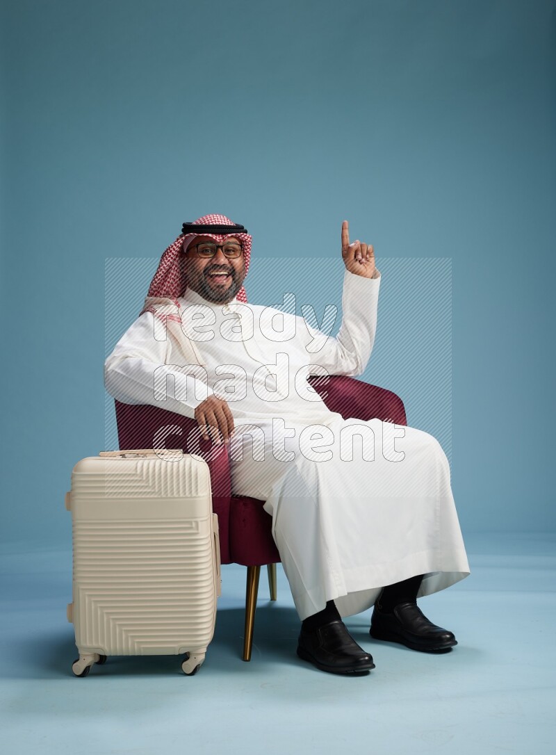 Saudi Man with shimag sitting on chair with a travel luggage on blue background