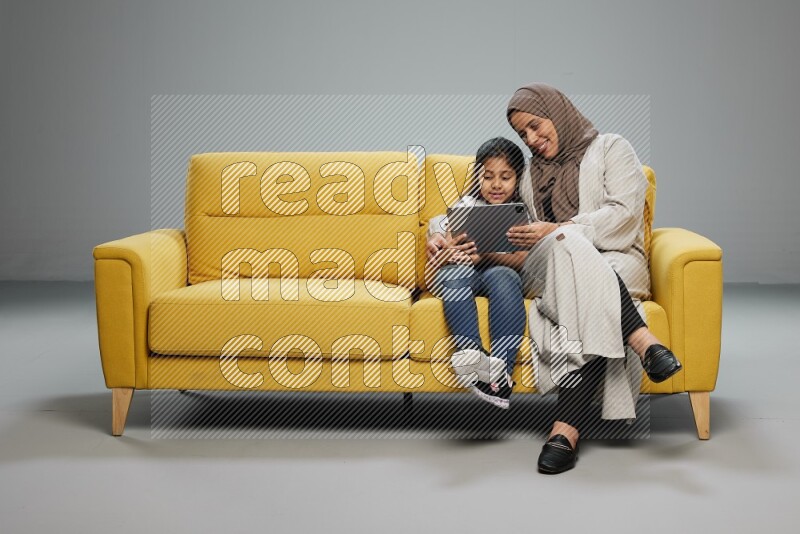 A girl with her mother on yellow sofa and watching on iPad on gray background