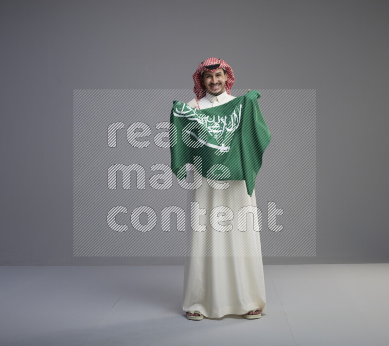 A saudi man standing wearing thob and red shomag holding big saudi flag on gray background