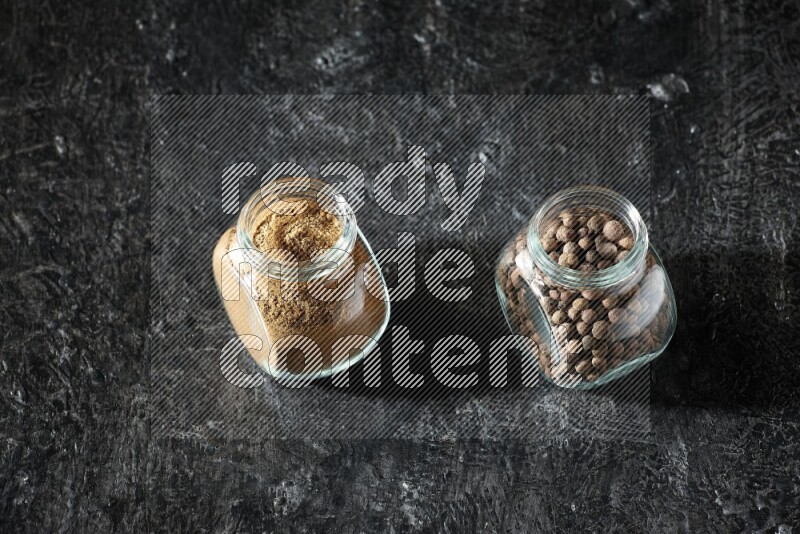 2 glass spice jars full of allspice powder and whole balls on a textured black flooring