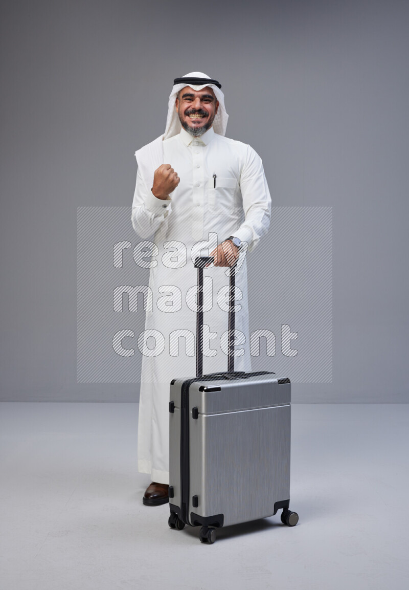 Saudi man wearing Thob and white Shomag standing holding Travel bag on Gray background